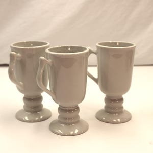 May include: Three light gray ceramic mugs with handles. The mugs have a pedestal base and a rounded body. The handles are curved and attached to the side of the mug. The mugs are arranged in a group.