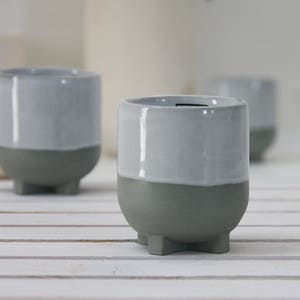 Op de afbeelding: Drie grijze keramische potten met een witte glazuur op de bovenste helft. De potten hebben een afgeronde vorm en drie kleine voetjes.