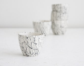 May include: A set of ceramic cups with a white base and a black splatter design. The cups have a rounded shape and are stacked in the background. The cups are suitable for serving beverages.