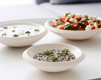 May include: Three white bowls on a white surface. The bowl on the left contains a white creamy sauce with green herbs. The bowl in the center contains a white and black mixture with green herbs. The bowl on the right contains a salad with tomatoes, cheese, and green herbs.