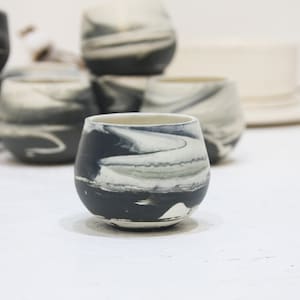 Puede incluir: Colección de copas de vino sin tallo de cerámica hechas a mano. Cada copa presenta un diseño veteado único en tonos grises y blancos, con una base gris oscuro. Las copas tienen forma redondeada y un acabado liso y brillante.