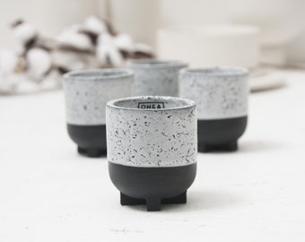 May include: Four small white ceramic cups with black bottoms and black feet. The cups have a speckled white glaze and the text "ONEA" is printed on the side of one cup.