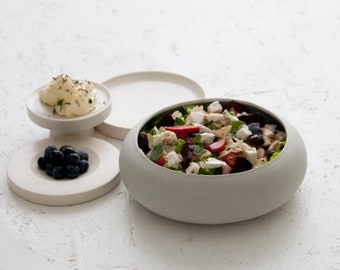 May include: A gray ceramic bowl filled with a salad of greens, feta cheese, strawberries, and beets. The bowl is on a white surface with two smaller gray ceramic dishes, one with blueberries and the other with a dollop of white cream.