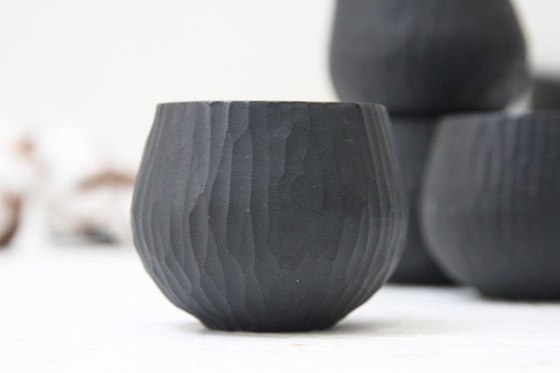 May include: Four black ceramic cups with a textured, wood-like finish. The cups are stacked on top of each other.