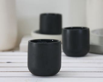Op de afbeelding: Drie zwarte keramische bekers, elk met een afgeronde bodem en een gladde, matte afwerking. De bekers staan op een wit oppervlak.