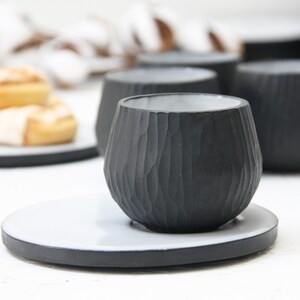 May include: A black ceramic cup with a textured exterior and a white saucer. The cup is sitting on a white surface.