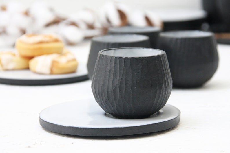 May include: A black ceramic cup with a textured exterior and a white saucer. The cup is sitting on a white surface.