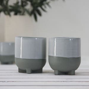 Op de afbeelding: Twee grijze keramische potten met een witte glazuur op de bovenste helft. De potten hebben drie kleine voetjes aan de onderkant.