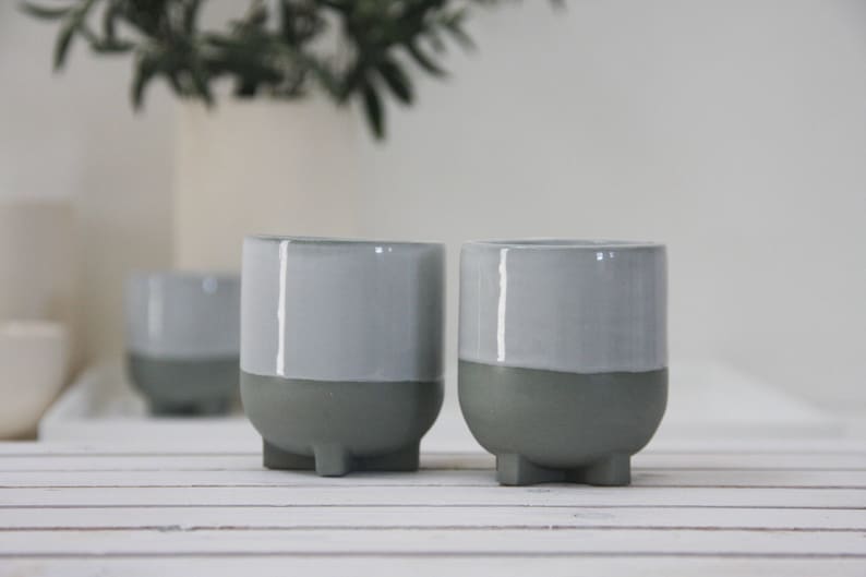 Op de afbeelding: Twee grijze keramische potten met een witte glazuur op de bovenste helft. De potten hebben drie kleine voetjes aan de onderkant.