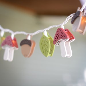 May include: A string of fairy lights with wooden cutouts of mushrooms, acorns, and leaves. The lights are white and the cutouts are painted in shades of red, brown, and green.