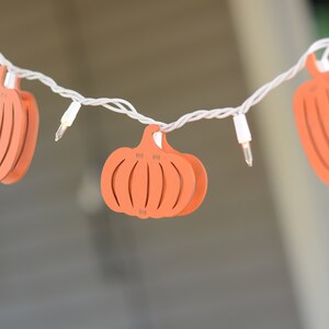 May include: A string of white fairy lights with orange pumpkin-shaped cut-outs. The pumpkins have a carved design and are strung together with white wire.