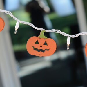 May include: A string of white lights with orange pumpkin-shaped cutouts. The pumpkins have a jack-o'-lantern face.