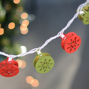Könnte beinhalten: Eine Lichterkette mit weißen Lichtern und roten und grünen Papierornamenten in Form von Schneeflocken. Die Ornamente sind mit Wäscheklammern an den Lichtern befestigt.