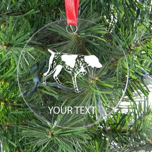 May include: Clear, round Christmas ornament with a white and black dog silhouette. The ornament hangs from a red ribbon and has the text "YOUR TEXT" at the bottom. The ornament is displayed against a backdrop of green Christmas tree branches.