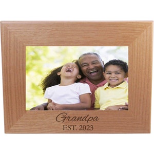 May include: A wooden picture frame with a photo of a grandfather and two grandchildren. The frame is engraved with the words "Grandpa EST. 2023".