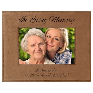 May include: A brown leatherette picture frame with the words "In Loving Memory" at the top. The frame holds a photo of two women. The bottom of the frame has the name "Batheny Kasso" and the dates 11.25.1934-07.23.2020.