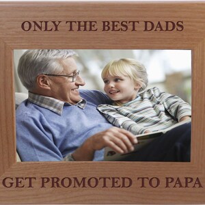 May include: A wooden picture frame with the text "Only the best dads get promoted to papa" engraved on the top and bottom. The frame contains a photo of an older man and a young girl smiling at each other.