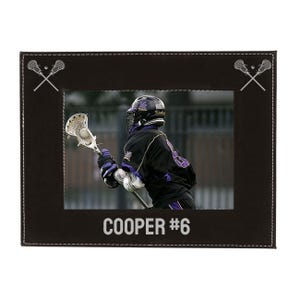 May include: A black leather picture frame with a stitched border. The frame has a silver lacrosse stick design in the top corners. The frame holds a photo of a lacrosse player wearing a black and purple uniform, holding a lacrosse stick. The text "COOPER #6" is printed below the photo.