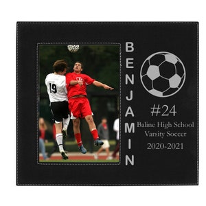 May include: Black leather picture frame with a photo of two soccer players in action. The frame has a silver inscription that reads "Benjamin #24 Baline High School Varsity Soccer 2020-2021" and a silver soccer ball graphic.