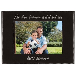 May include: A black leather photo frame with a white stitched border. The frame has a photo of a father and son holding a soccer ball. The text "The love between a dad and son lasts forever" is inscribed on the frame.