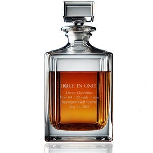 May include: A clear glass decanter with a silver stopper. The decanter is filled with a dark amber liquid. The decanter is engraved with the text "HOLE IN ONE! Danny Ganderine Hole #4 122 yards 7 iron Sweetgum Golf Course May 14, 2023".