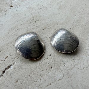 May include: Two silver-colored clam shell-shaped objects. The shells have a textured surface with visible ridges. The objects are displayed on a light-colored, textured surface.