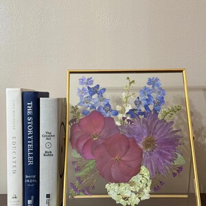 May include: A gold framed glass display with a pressed flower arrangement. The flowers are purple, blue, and white. The frame is sitting on a wooden surface with three books.