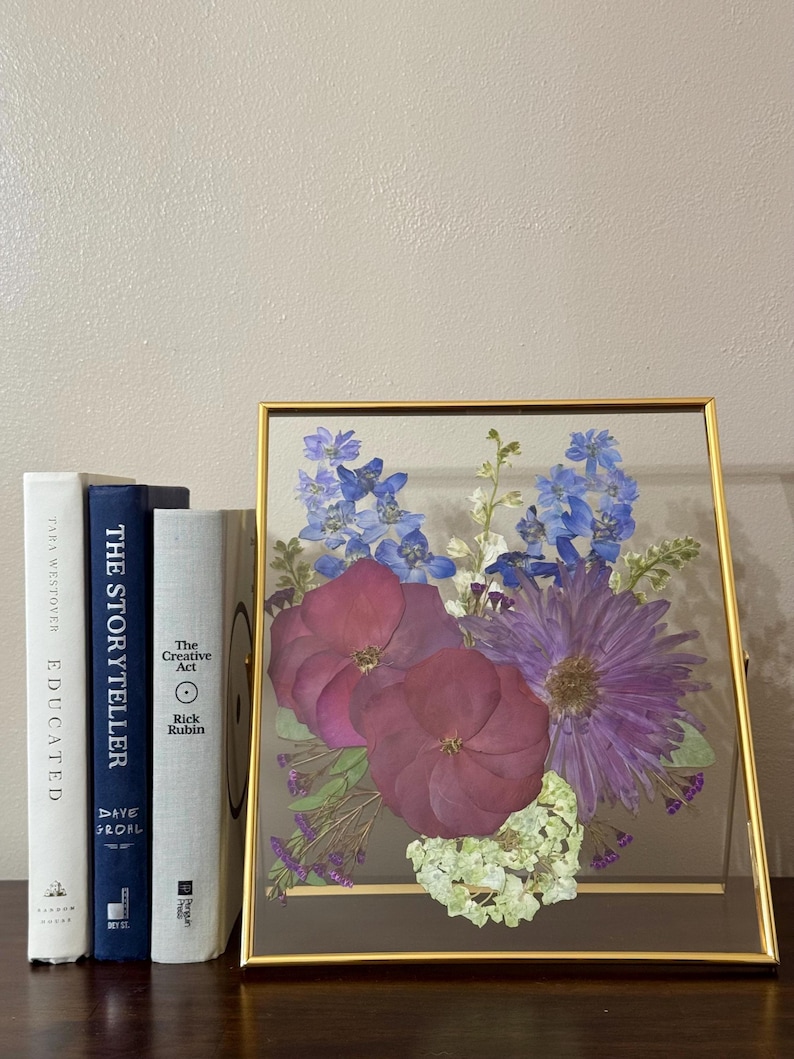 May include: A gold framed glass display with a pressed flower arrangement. The flowers are purple, blue, and white. The frame is sitting on a wooden surface with three books.
