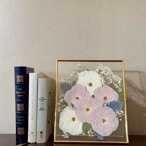 May include: A gold framed picture with a pressed flower arrangement. The flowers are white and light purple and are arranged in a bouquet. The frame is on a wooden surface.