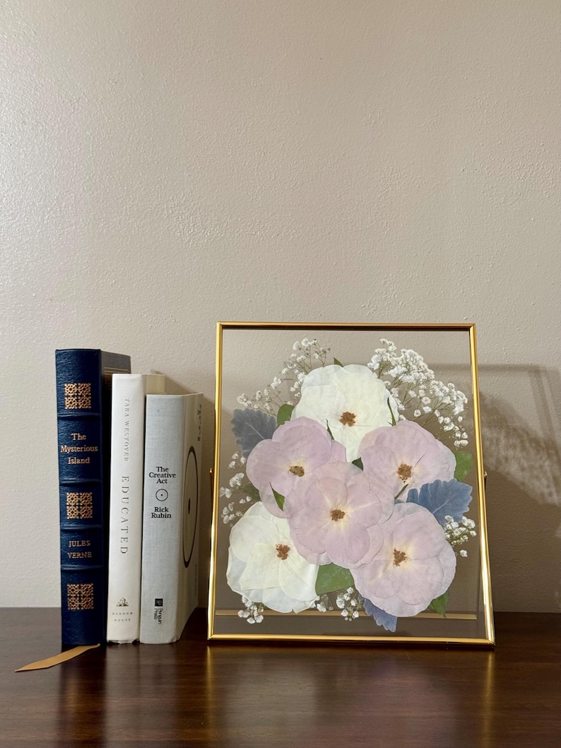 May include: A gold framed picture with a pressed flower arrangement. The flowers are white and light purple and are arranged in a bouquet. The frame is on a wooden surface.