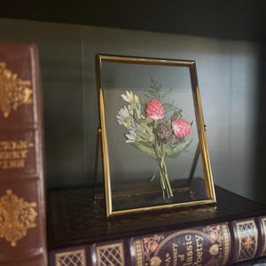 May include: A gold-framed picture with a pressed flower arrangement inside. The flowers are pink and white and are arranged in a bouquet. The picture is sitting on a stack of books.