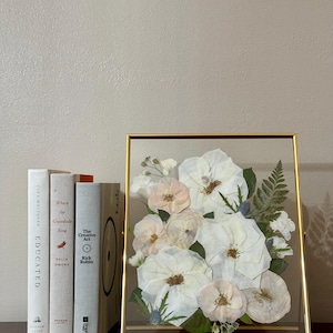 May include: A framed pressed flower arrangement with white and pink roses, ferns, and other greenery. The frame is gold and has a glass front.