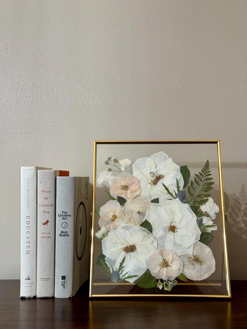 May include: A framed pressed flower arrangement with white and pink roses, ferns, and other greenery. The frame is gold and has a glass front.