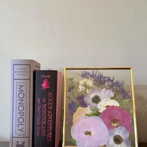 May include: A gold framed glass display with pressed flowers. The flowers are shades of pink, white, and purple. The frame is sitting on a table next to two books. One book is a vintage edition of Monopoly and the other is a copy of Alice's Adventures in Wonderland by Carroll.
