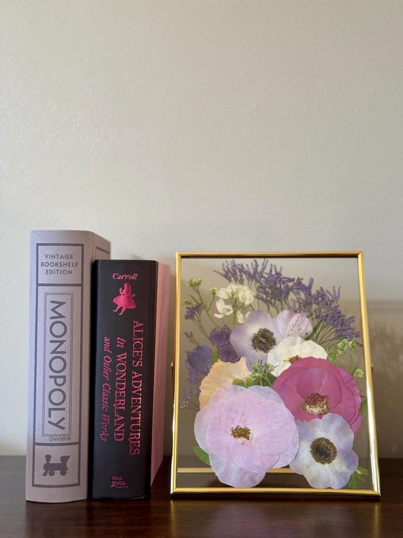 May include: A gold framed glass display with pressed flowers. The flowers are shades of pink, white, and purple. The frame is sitting on a table next to two books. One book is a vintage edition of Monopoly and the other is a copy of Alice's Adventures in Wonderland by Carroll.
