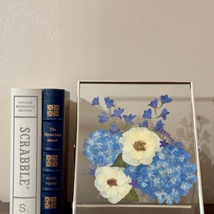 May include: A framed pressed flower arrangement with white and blue flowers. The flowers are arranged in a cluster and are pressed between two pieces of glass. The frame is silver and has a simple design.