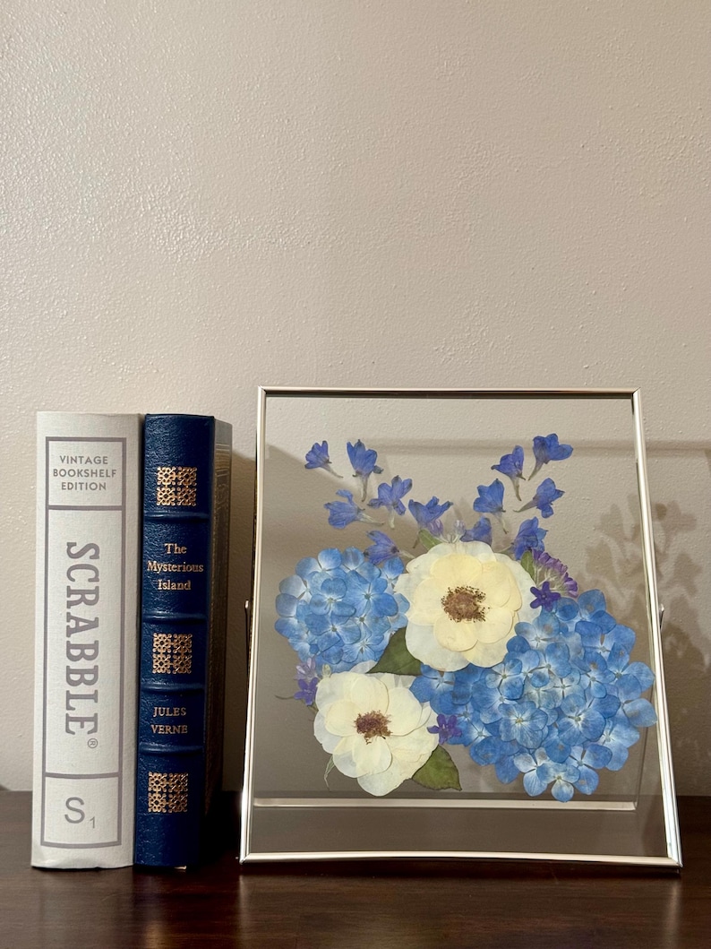 May include: A framed pressed flower arrangement with white and blue flowers. The flowers are arranged in a cluster and are pressed between two pieces of glass. The frame is silver and has a simple design.