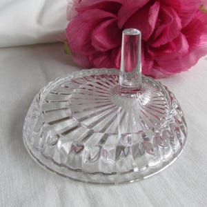 Könnte beinhalten: Ein klarer Glas-Ringhalter mit rundem, geriffeltem Sockel und vertikalem Pfosten. Der Ringhalter steht vor einem weißen Hintergrund mit einer rosa Blume im Hintergrund. Der Ringhalter ist ein dekorativer Gegenstand.