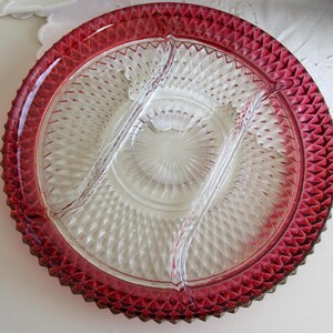 May include: A round, divided glass serving dish featuring a clear center with a sunburst pattern and a deep red, diamond-cut textured rim. The dish is partitioned into three sections by two curved, clear glass dividers that extend across the surface.
