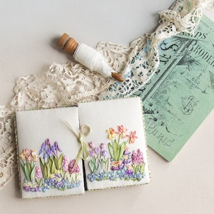 Puede incluir: Dos libros blancos cubiertos de tela con bordados de cintas coloridas de flores de primavera, incluyendo iris, narcisos e jacintos. También hay una bobina de madera envuelta en encaje blanco y un libro verde vintage con texto en francés. Los libros están atados con una cinta verde.