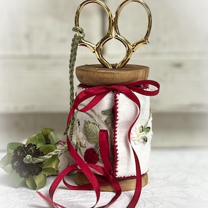 Puede incluir: Una bobina decorativa con tijeras doradas, cinta roja y bordados florales. La bobina es de madera y está envuelta en tela blanca con flores bordadas y detalles rojos. Una cuerda verde está unida a las tijeras.