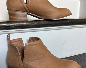 May include: A pair of tan leather ankle boots with a side cut-out design. The boots have a low, stacked heel and a rounded toe. The stitching is visible along the sole. The boots are displayed on a staircase.