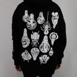 May include: Black hoodie with a white graphic print of light bulbs and insects. The light bulbs have scenes inside them, such as a beach, a forest, and a campfire.