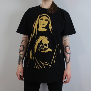 May include: Black t-shirt with a gold print of a woman holding a skull. The woman is wearing a veil and has tears on her face. The text "Live" and "Not Life" is printed on the sleeves.