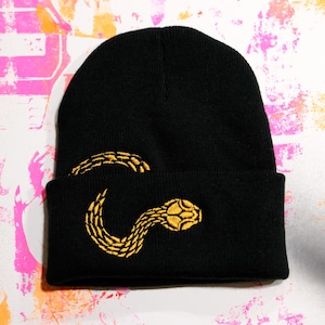 May include: Black knit beanie with a gold embroidered snake design. The snake is coiled and detailed, adding a unique touch to the hat. A stylish and warm accessory for any wardrobe.