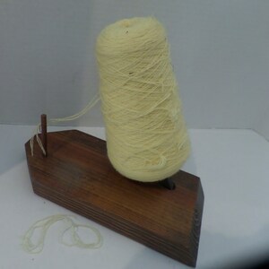May include: A wooden yarn holder with a skein of pale yellow yarn. The yarn is wound around a central spindle and the holder has a small wooden peg on the side.