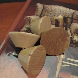 May include: Four wooden knobs with a natural finish. The knobs are round with a slight cone shape and have a screw on the bottom. The knobs are sitting on a brown book.