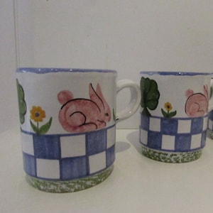 May include: Three ceramic mugs with a blue and white checkered pattern, green leaves, and pink bunny illustrations. Each mug has a handle.