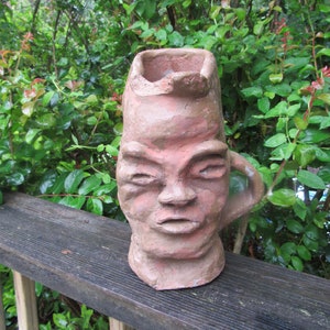 May include: A terracotta sculpture of a face with a wide mouth and closed eyes. The sculpture is in the shape of a pitcher with a handle.