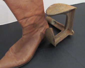 May include: A brown leather boot with a pointed toe is placed on a metal boot jack. The boot jack is rusty and has a curved shape.